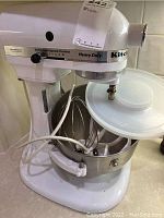 Left side profile of white KitchenAid Heavy Duty mixer showing speed control and mixing bowl with plastic cover and attachments inside bowl.