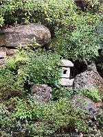 Small square multi-tier cement pagoda lantern partially obscured by green plants and rocks among garden landscaping.