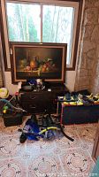 Wide view showing multiple swimming flippers, helmets, and assorted sports gear around a small chest of drawers with a framed painting behind.