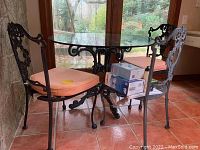 Front-left side view of the round glass top table with three matching cast iron chairs, two with salmon cushions.