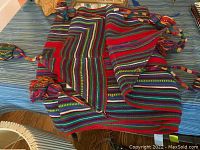 Table runner folded showing vibrant multicolor stripes, mainly red, blue, green, and gold with braided tassels at the ends.