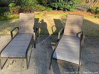 Pair of outdoor lounge chairs pictured on a concrete patio and grassy lawn in daylight. Both chairs have bronze colored metal frames and light tan fabric seats and backs with a woven design. The chairs have armrests and adjustable reclining backs.