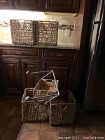 Five wicker storage baskets in various sizes placed on floor and countertop in kitchen setting.