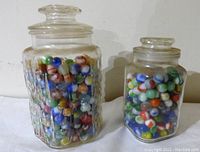 Two glass jars, one large and one smaller, filled with mixed colorful glass marbles.