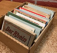 Photo of a box filled with assorted vintage postcard sets showing the top portions of various envelopes and brochures, with one titled 'Der Rhein' in front