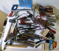 Overview of assorted vintage hand tools laid out on a flat surface showing saws, hammers, knives, blades, crowbar, mallet, chisels, and other tools