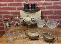 Full lot view showing all six sets of creamer and sugar bowls arranged with some on elevated clear stands and a vintage Vogue Kroc box behind.