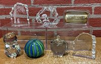 Seven paperweights displayed on clear stands in front of a brick wall, including shaped glass pieces and a photo embedded glass paperweight.