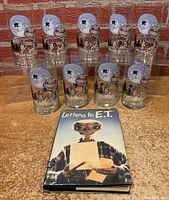 Nine glass tumblers arranged in two rows with colorful E.T. graphics and 'Phone home' text, plus 'Letters to E.T.' book with dust jacket laid in front