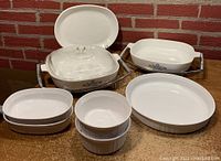 Photo showing 8 pieces of CorningWare including round and oval casseroles, serving platter, quiche dish, skillets with lid and metal cradle on a countertop against brick wall background.