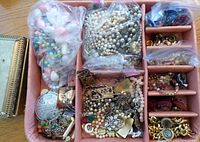 Top view of pink jewelry box filled with multiple zip-lock bags holding assorted vintage costume jewelry components, plus visible small pastel faux pearl necklaces, metal chains, and various charms. Jewelry items appear used with visible wear.
