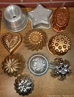 Nine vintage tin and metal molds and pans arranged on a surface, showing shapes such as star, heart, floral, and round forms, in copper, aluminum, and tin material.