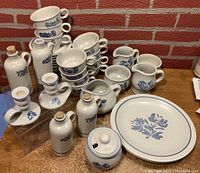 Photo showing complete set of white ceramic dinnerware and accessories with blue floral pattern, including plates, cups, saucers, creamers, sugar bowls, candle holders, vinegar and oil bottles, jam/jelly jar, and gravy boat arranged on counter against brick wall.