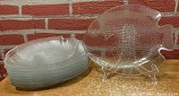 Stack of nine clear fish-shaped glass plates next to one plate displayed upright on a stand to show shape and texture details.