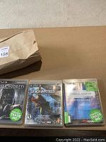 Flat lay photo of three PS3 game cases: Dishonored, Uncharted 2 Among Thieves, and Hitman Absolution on a brown surface with a paper bag to the top left.