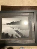 Black and white print showing a rowboat docked near grassy shore and misty lake with dark tree line in background