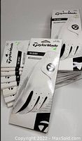 Nine packaged and loose TaylorMade sport golf gloves arranged for viewing; white gloves with black holes and branding visible, mostly new in box.