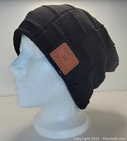 Black knit floppy beanie hat with quilt pattern displayed on mannequin head. Leather patch on side has perforations for embedded Bluetooth speaker.