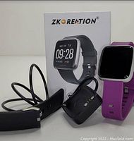 Image showing ZKOREATION smartwatch with purple band on the device, a black band detached, magnetic charger, and the product box in the background with branding and display image.