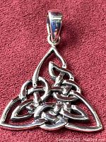 Front view of the Celtic triquetra knot pendant on a reddish fabric background, showing intricate sterling silver knot design and bail for chain attachment.