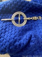 Full view of the Celtic pewter scarf pin lying on a blue textured fabric, showing the circular knot ring and long decorative pin.