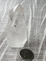 Clear quartz crystal point standing next to a Canadian dime for size reference, showing the polished six-sided prism shape and visible phantom ghost inclusions inside.