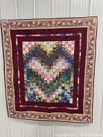 Full view of handmade quilt featuring a multi-colored heart design with burgundy borders and floral trim hung against a wall.