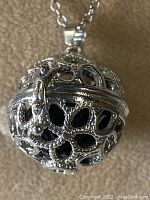 Close-up photo of the silver-tone ornate spherical locket pendant revealing intricate filigree design and black lava stone inside.