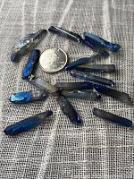 Top-down view of multiple mystic quartz points scattered with a coin for size reference, showing metallic blue-purple iridescence and drilled holes for jewelry