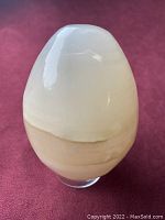 Front view of the hand polished onyx egg showing creamy white and beige layered stone with irregular shape, sitting on a clear plastic stand.