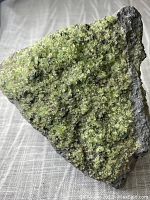 Front view of the mineral specimen showing bright green peridot crystals densely covering the triangular surface of the gray rock matrix on fabric background.