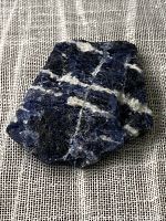 Photo showing one side of a single rough sodalite mineral stone with rich blue color and white streaks on a textured fabric background.