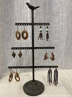 Full view of metal bird earring stand with six pairs of pierced earrings displayed on it, showing a variety of wooden, beaded, and leather earrings.