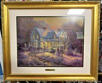 Framed artwork showing a winter house scene with snow and a snowman, in gold frame.