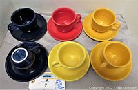Six Fiesta ware cups and saucers in various colors including blue, red, and yellow arranged on a surface.
