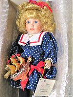 Porcelain doll with blonde hair wearing navy dress with white stars and red bows, holding a small ship model, displayed in box.