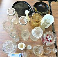 Photo showing a collection of clear and amber ball jars, vases in white, yellow, and clear glass, two black glass dishes, a glass bell, and small decorative glassware items on a wood grain surface.