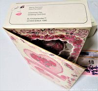 Box opened showing doll inside and design resembling a Valentine's Day greeting card with floral and heart patterns, classic decorative style of pink, red and white colors.