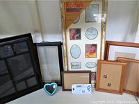 Box lot of multiple photo frames including a Kodak collage, wooden frames in various sizes, and a small blue heart-shaped frame.