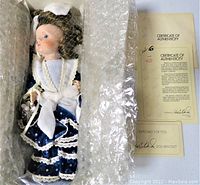 Porcelain doll inside box wrapped partially in bubble wrap, visible certificate of authenticity and doll bracelet included.