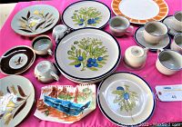 Overview of various stoneware plates, cups, and platter with colorful botanical patterns including blue berries, green leaves, orange geometric, and brown wheat designs on pink cloth background.