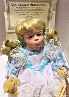 Frontal view of Vintage Tearful Tammy doll seated with Certificate of Authenticity behind. Doll has blonde hair in pigtails tied with blue and yellow ribbons, blue floral dress with white apron and lace. Small brown teddy bear held in left hand.