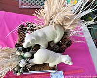 Overview of two white polar bear figurines on silver box with pinecones and white dried plants on pink tablecloth.