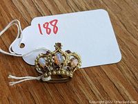 Gold-tone crown brooch with simulated opal stones and purple rhinestones on a wooden surface with a tag labeled 188.