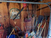 Photo showing collection of long handled rakes, shovels, and other yard tools leaning against wooden wall