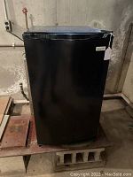 Front view photo of black Magic Chef mini refrigerator placed on a platform in a basement or storage area.