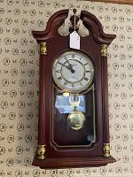 Full view of modern colonial style wall clock with wooden frame and brass accents, glass pendulum window visible.