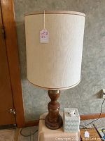 Side view of one vintage table lamp on small stand showing turned wooden base, brass accents, and cream fabric shade with brown trim.