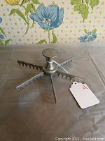 Metal pie slicing guide placed on a table with floral wallpaper background. Shows the tool from a low side angle highlighting the knob and slicing blades.