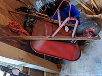 Small red steel wheelbarrow shown lying on its side with wooden handles and one rubber wheel, placed on a floor with gardening tools nearby.